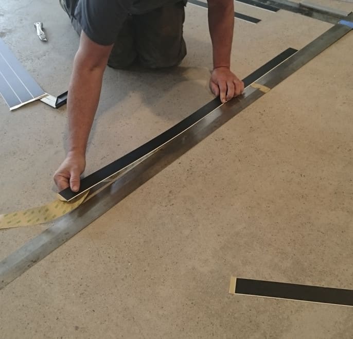 Installation av akryl-ledstråk. Bilden visar hur man drar av skyddspappret på stråkets baksida, sen riktar in rätt placering med hjälp av en lång stållinjal utplacerad på golvet. Därefter fästs stråket mot det rengjorda golvet.