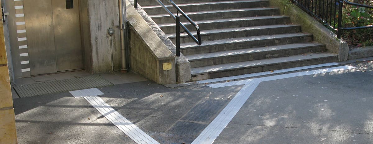 Taktila ledstråk av gummi installerade i utomhusmiljö på Herrljunga station. Ljusgrått ledstråk som leder fram till uppmarksamhetsmarkeringar vid hiss till vänster och trappa till höger.