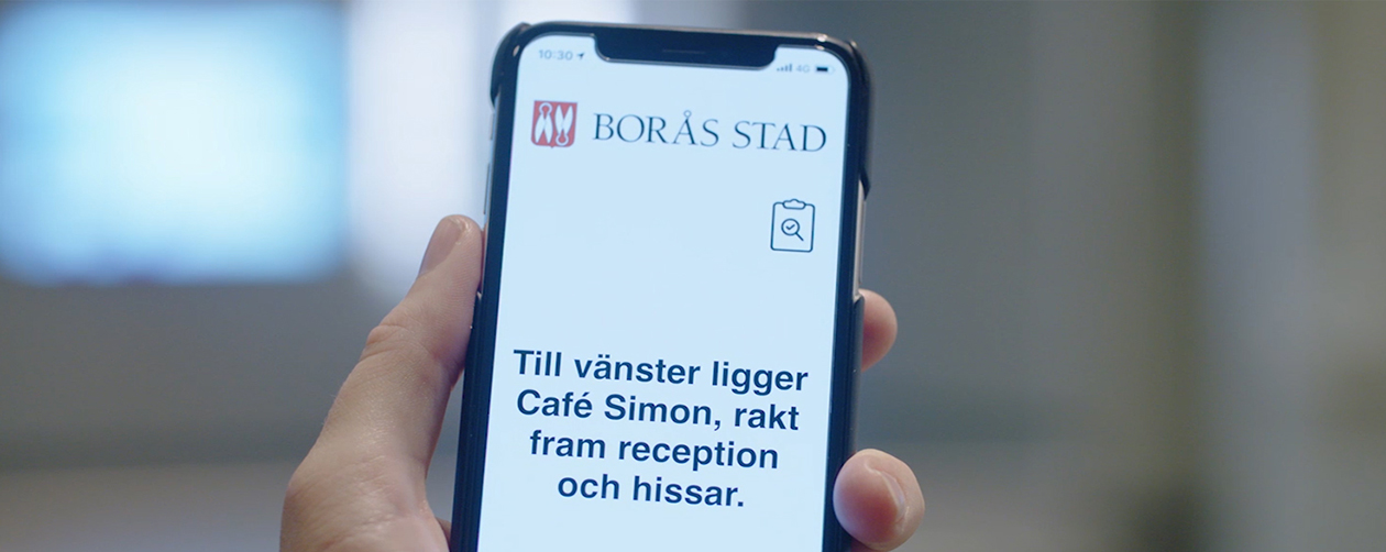 Tactile Flooring App - A smarter wayfinding. Bilden visar en hand med en mobiltelefon. I fönstret på mobilen visas en informationstext: Till vänster ligger Café Simon, rakt fram reception och hissar.