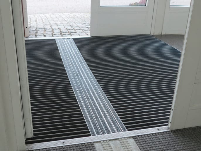 Vår entré med Aluminiumprofilmatta med taktilt ledstråk ATF 122 med inlägg i ljusgrå nålfilt. Mattan vid sidan av stråket är med svart gummi-inlägg.