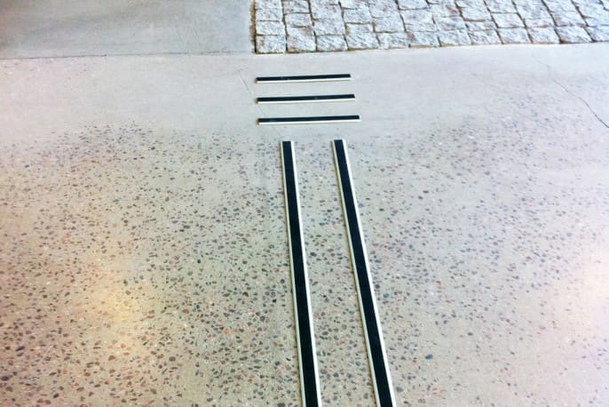 Bilden visar 2 aluminium ledstråk (38 mm) som leder fram till ett "stopp" som är markerat med 3 tvärställda ca 30 cm långa profiler. de ligger på ett betonggolv och i överkant synas en yta med gatsten. Bilden är fotad på Simonsland i Borås.