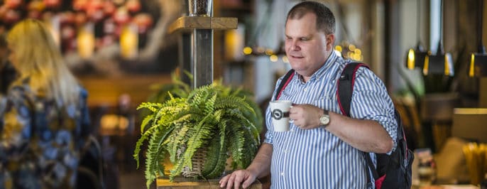 Fredrik dricker kaffe, ståendes vid ett bord på kafé. Bredvid på bänken står en vacker grön ormbunke.