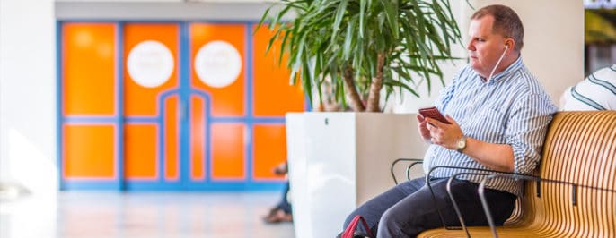 Bild från ett resecentrum där Fredrik sitter på en soffa, i handen håller han sin mobil. I bakgrunden skymtar en kruka med ett träd och där bakom en dörr i vackert orange och blått.