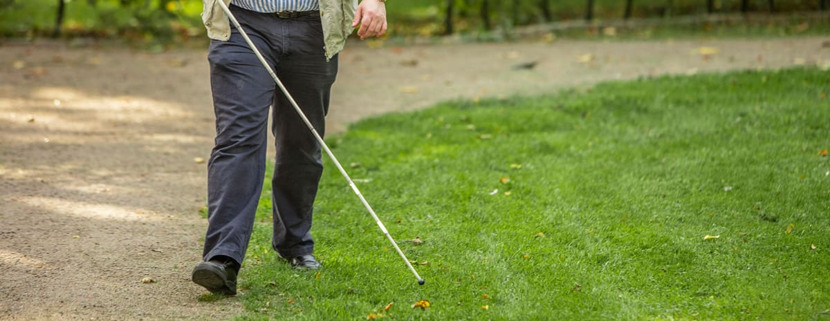 Klippt bild som visar underkroppen på Fredrik som promenerar i en park med vit käpp. Han använder gräskanten intill grusgången som ledstråk/hållpunkt.