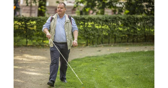 Helbild på Fredrik som promenerar i en park med vit käpp. Han använder gräskanten intill grusgången som ledstråk/hållpunkt.
