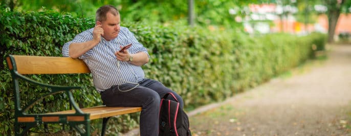Fredrik sitter på en parkbänk, i handen har han sin mobil och en hörsnäcka i örat.